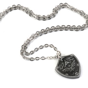 May include: A silver chain necklace with a pendant shaped like a shield with a stylized design of a bird and a triangle. The pendant is dark gray and has a textured surface.