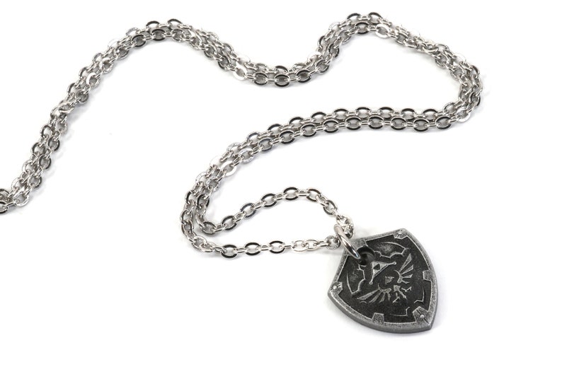 May include: A silver chain necklace with a pendant shaped like a shield with a stylized design of a bird and a triangle. The pendant is dark gray and has a textured surface.