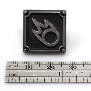 K&ouml;nnte beinhalten: Eine silberne Metallstifte mit einem schwarzen quadratischen Rahmen und einem stilisierten Flammenmuster im Inneren. Die Flamme ist ein Kreis mit einer Flamme, die aus der Oberseite herausragt.