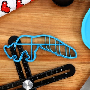 May include: A blue plastic cookie cutter shaped like a cartoon animal, possibly a fox or a raccoon, on a wooden surface with a ruler and a blue spatula.