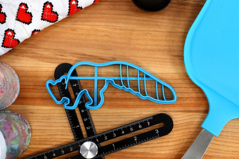 May include: A blue plastic cookie cutter shaped like a cartoon animal, possibly a fox or a raccoon, on a wooden surface with a ruler and a blue spatula.