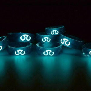 May include: A stack of glow-in-the-dark silicone rings with a white symbol on each ring. The rings are arranged in a pyramid shape and are glowing a bright teal color.