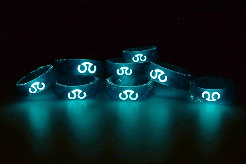 May include: A stack of glow-in-the-dark silicone rings with a white symbol on each ring. The rings are arranged in a pyramid shape and are glowing a bright teal color.