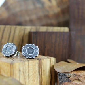 May include: A pair of silver-colored metal earrings with a circular design. The earrings are on a wooden surface.