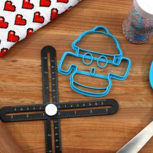 May include: A blue plastic cookie cutter shaped like a snowman with a hat, eyes, and a mouth. The cutter is on a wooden surface with a black ruler, a rolling pin, and a blue spatula.