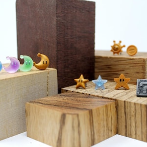 May include: A set of colourful and metallic earrings featuring various video game characters and items, including a gold coin, a star, a question mark block, and a mushroom. The earrings are displayed on wooden blocks.