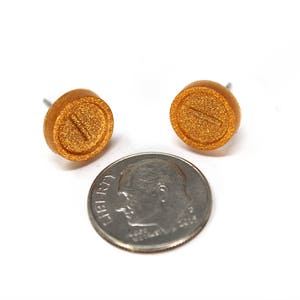 May include: A pair of gold-colored stud earrings with a pill shape and a line down the center. The earrings are about the size of a dime.