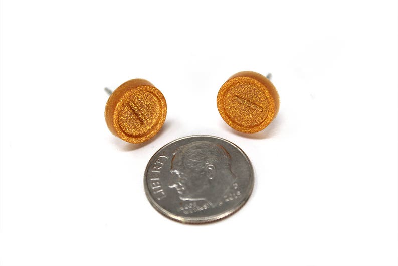 May include: A pair of gold-colored stud earrings with a pill shape and a line down the center. The earrings are about the size of a dime.