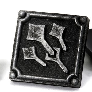 May include: A black and silver metal pin with a stylized design. The pin is square shaped and has a pin back.