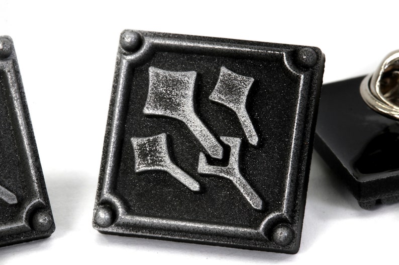 May include: A black and silver metal pin with a stylized design. The pin is square shaped and has a pin back.
