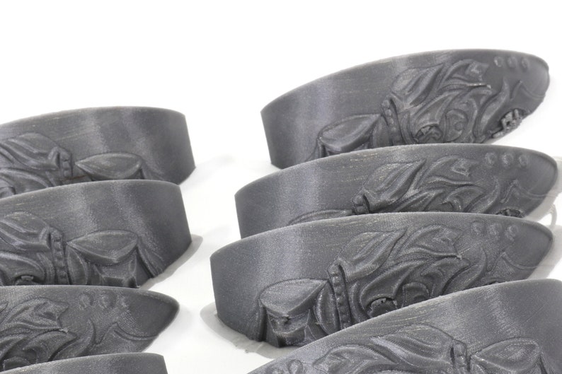 May include: A set of eight grey 3D printed decorative pieces with an intricate design. The pieces are shaped like a curved, flat surface with a raised design.