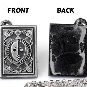 Puede incluir: Un collar de plata con un colgante que presenta una cara en blanco y negro con un dise&ntilde;o de sol. La parte posterior del colgante es lisa y negra. Las palabras "FRONT" y "BACK" est&aacute;n impresas sobre el colgante.