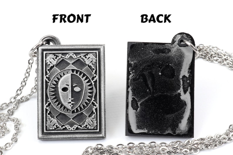 Puede incluir: Un collar de plata con un colgante que presenta una cara en blanco y negro con un dise&ntilde;o de sol. La parte posterior del colgante es lisa y negra. Las palabras "FRONT" y "BACK" est&aacute;n impresas sobre el colgante.