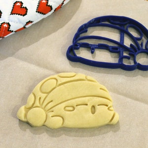 Pode incluir: Um cortador de biscoitos de pl&aacute;stico azul e um biscoito marrom claro em forma de personagem de desenho animado. O cortador de biscoitos e o biscoito est&atilde;o em uma superf&iacute;cie de papel marrom.
