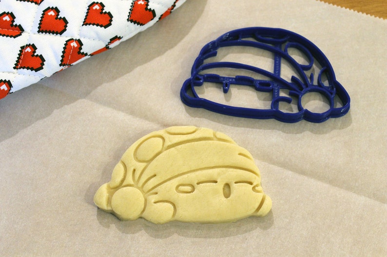 Pode incluir: Um cortador de biscoitos de pl&aacute;stico azul e um biscoito marrom claro em forma de personagem de desenho animado. O cortador de biscoitos e o biscoito est&atilde;o em uma superf&iacute;cie de papel marrom.