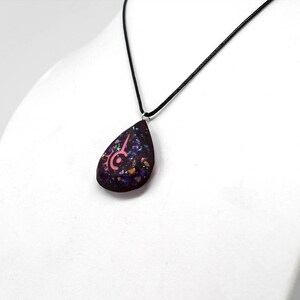 May include: A teardrop-shaped pendant necklace with a black cord. The pendant is dark purple with iridescent flecks and a pink design. The necklace is displayed on a white bust.