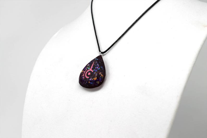 May include: A teardrop-shaped pendant necklace with a black cord. The pendant is dark purple with iridescent flecks and a pink design. The necklace is displayed on a white bust.