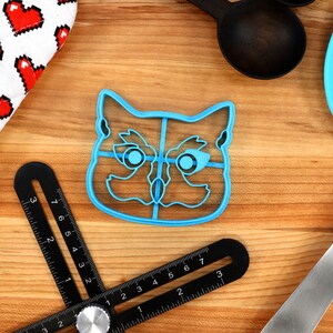May include: Blue plastic cat-shaped cookie cutter with a divided interior. The cutter is on a wooden surface.