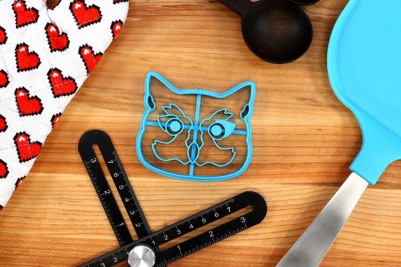 May include: Blue plastic cat-shaped cookie cutter with a divided interior. The cutter is on a wooden surface.