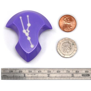 May include: A purple, irregularly shaped dice with a white constellation design. The dice is about the size of a US penny.