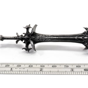 May include: A silver-colored sword with a detailed, intricate design. The sword is approximately 9.5 inches long and has a sharp point. The sword is made of metal and has a textured surface.