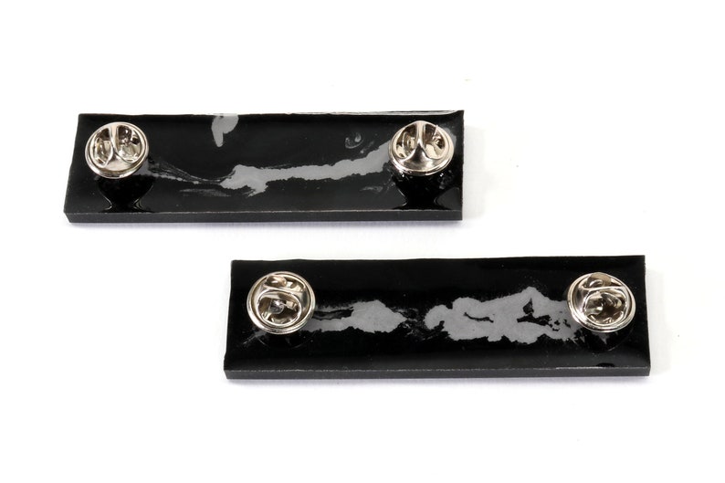 May include: Two black enamel pins with silver metal backs. The pins are rectangular and have a white design on them.