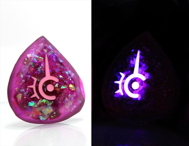 May include: A teardrop-shaped, pink resin pendant with a glowing white symbol. The pendant is filled with iridescent glitter and has a white symbol on the front. The pendant glows purple in the dark.