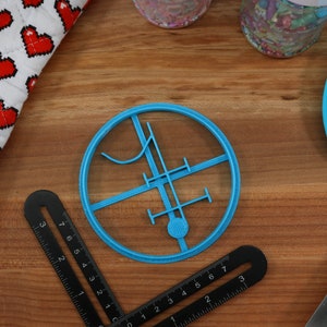 Op de afbeelding: Een blauwe plastic koekjesvormer in de vorm van een cirkel met een ontwerp erin. Het ontwerp bevat een verticale lijn, een horizontale lijn en een gebogen lijn.