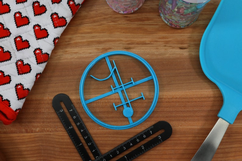Op de afbeelding: Een blauwe plastic koekjesvormer in de vorm van een cirkel met een ontwerp erin. Het ontwerp bevat een verticale lijn, een horizontale lijn en een gebogen lijn.