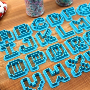 May include: A set of blue plastic cookie cutters shaped like the letters of the alphabet in a pixelated style. The cutters are arranged on a wooden surface.