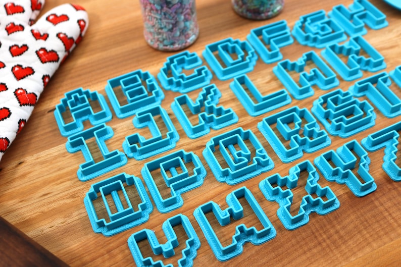 May include: A set of blue plastic cookie cutters shaped like the letters of the alphabet in a pixelated style. The cutters are arranged on a wooden surface.