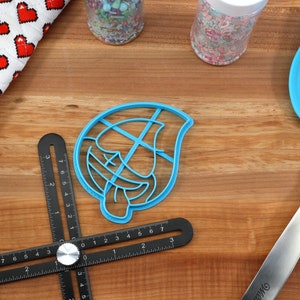 May include: A blue plastic cookie cutter shaped like a cartoon face with a tongue sticking out. The cutter is on a wooden surface with a black ruler, a rolling pin, and a blue spatula.