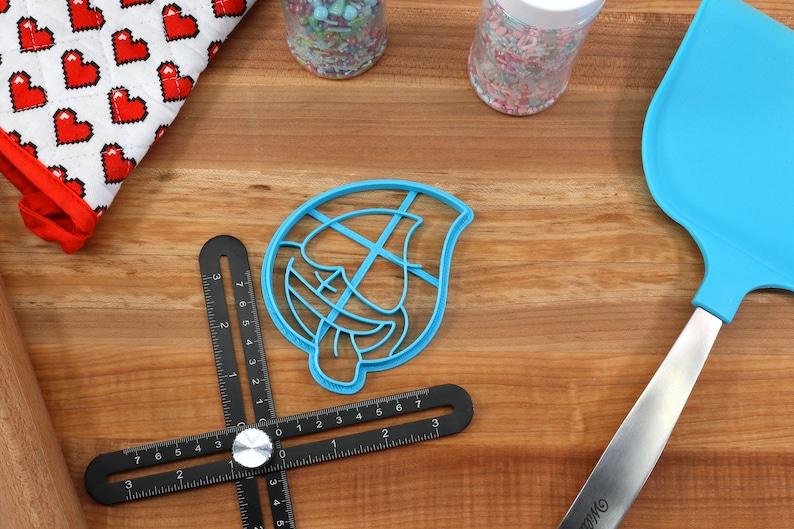 May include: A blue plastic cookie cutter shaped like a cartoon face with a tongue sticking out. The cutter is on a wooden surface with a black ruler, a rolling pin, and a blue spatula.