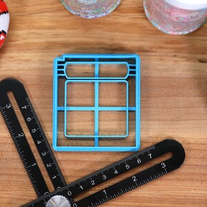 May include: A blue plastic cookie cutter shaped like a window with six panes. The cutter is on a wooden surface with a ruler and other items.