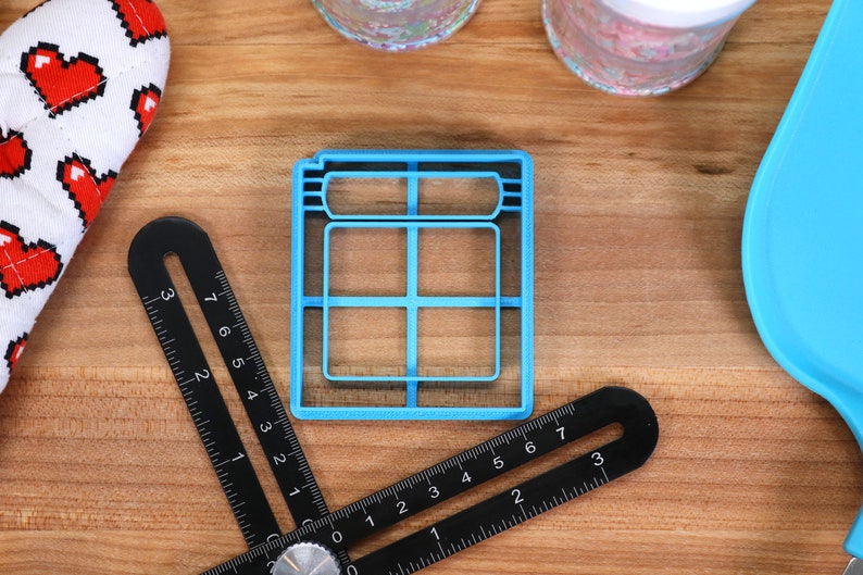 May include: A blue plastic cookie cutter shaped like a window with six panes. The cutter is on a wooden surface with a ruler and other items.