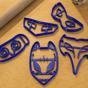 May include: A collection of blue plastic cookie cutters in various shapes, including a mask, a fox, and abstract designs, are arranged on a wooden surface. A wooden rolling pin and a heart-patterned oven mitt are also visible.