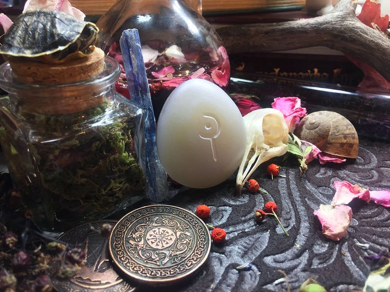 May include: A white egg-shaped stone with a symbol carved into it, surrounded by a variety of objects including a small turtle, a glass jar with moss, a blue crystal, a skull, a snail shell, and pink flower petals on a dark patterned surface.