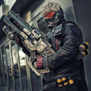May include: A cosplayer dressed as a video game character, wearing a black leather jacket, a red and black helmet, and holding a large, futuristic-looking gun.