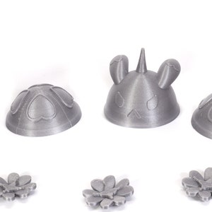 May include: Four 3D printed gray plastic figurines. Two are shaped like a dome with hearts on the surface. One is shaped like a dome with a unicorn head on top. The last is shaped like a dome with bunny ears on top. Three gray plastic flowers are also shown.