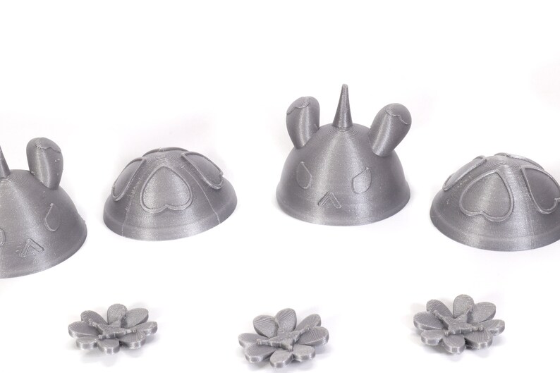 May include: Four 3D printed gray plastic figurines. Two are shaped like a dome with hearts on the surface. One is shaped like a dome with a unicorn head on top. The last is shaped like a dome with bunny ears on top. Three gray plastic flowers are also shown.
