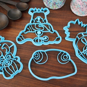 May include: Four blue plastic cookie cutters shaped like a snowman, a pineapple, a fish, and a face with a beard. The cutters are on a wooden surface.