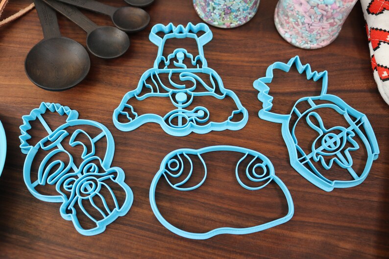 May include: Four blue plastic cookie cutters shaped like a snowman, a pineapple, a fish, and a face with a beard. The cutters are on a wooden surface.