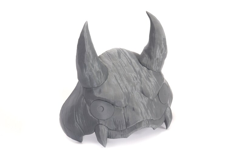 May include: A gray, textured, 3D printed mask with horns and a detailed design. The mask has a textured surface that resembles wood grain.