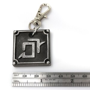 K&ouml;nnte beinhalten: Ein silberner Schl&uuml;sselanh&auml;nger mit einem quadratischen Anh&auml;nger, der ein stilisiertes Symbol zeigt. Der Anh&auml;nger hat einen schwarzen Hintergrund und ein silbernes Symbol. Der Schl&uuml;sselanh&auml;nger ist an einem silbernen Ring und einem silbernen Verschluss befestigt.