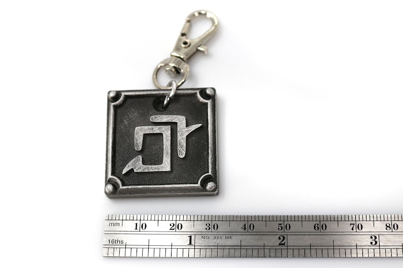K&ouml;nnte beinhalten: Ein silberner Schl&uuml;sselanh&auml;nger mit einem quadratischen Anh&auml;nger, der ein stilisiertes Symbol zeigt. Der Anh&auml;nger hat einen schwarzen Hintergrund und ein silbernes Symbol. Der Schl&uuml;sselanh&auml;nger ist an einem silbernen Ring und einem silbernen Verschluss befestigt.