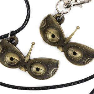 May include: Two gold-colored robot eye charms, one on a black cord and the other on a silver key chain. Each charm features a large eye with a small antenna.