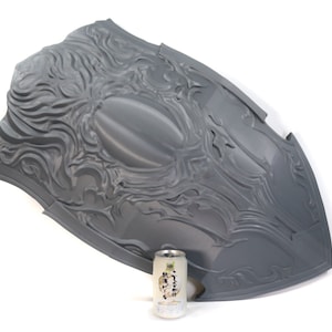 May include: A large, gray, plastic shield with a detailed, embossed design. The shield is shaped like a traditional medieval shield and has a raised, oval center. The shield is approximately 30 inches in diameter.