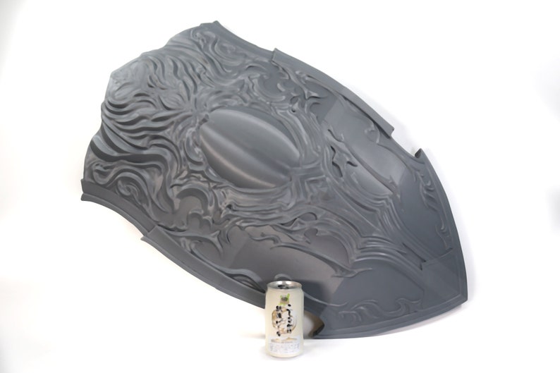 May include: A large, gray, plastic shield with a detailed, embossed design. The shield is shaped like a traditional medieval shield and has a raised, oval center. The shield is approximately 30 inches in diameter.