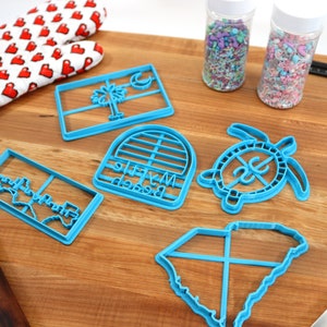 May include: A set of six blue plastic cookie cutters featuring various designs, including a state outline, a turtle, a palm tree, and a beach-themed arch. The cutters are arranged on a wooden surface with sprinkles in jars and a rolling pin.