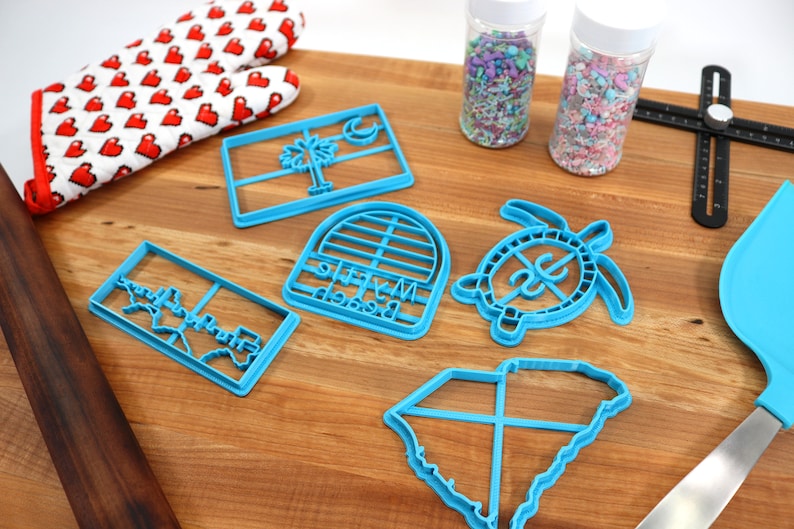 May include: A set of six blue plastic cookie cutters featuring various designs, including a state outline, a turtle, a palm tree, and a beach-themed arch. The cutters are arranged on a wooden surface with sprinkles in jars and a rolling pin.